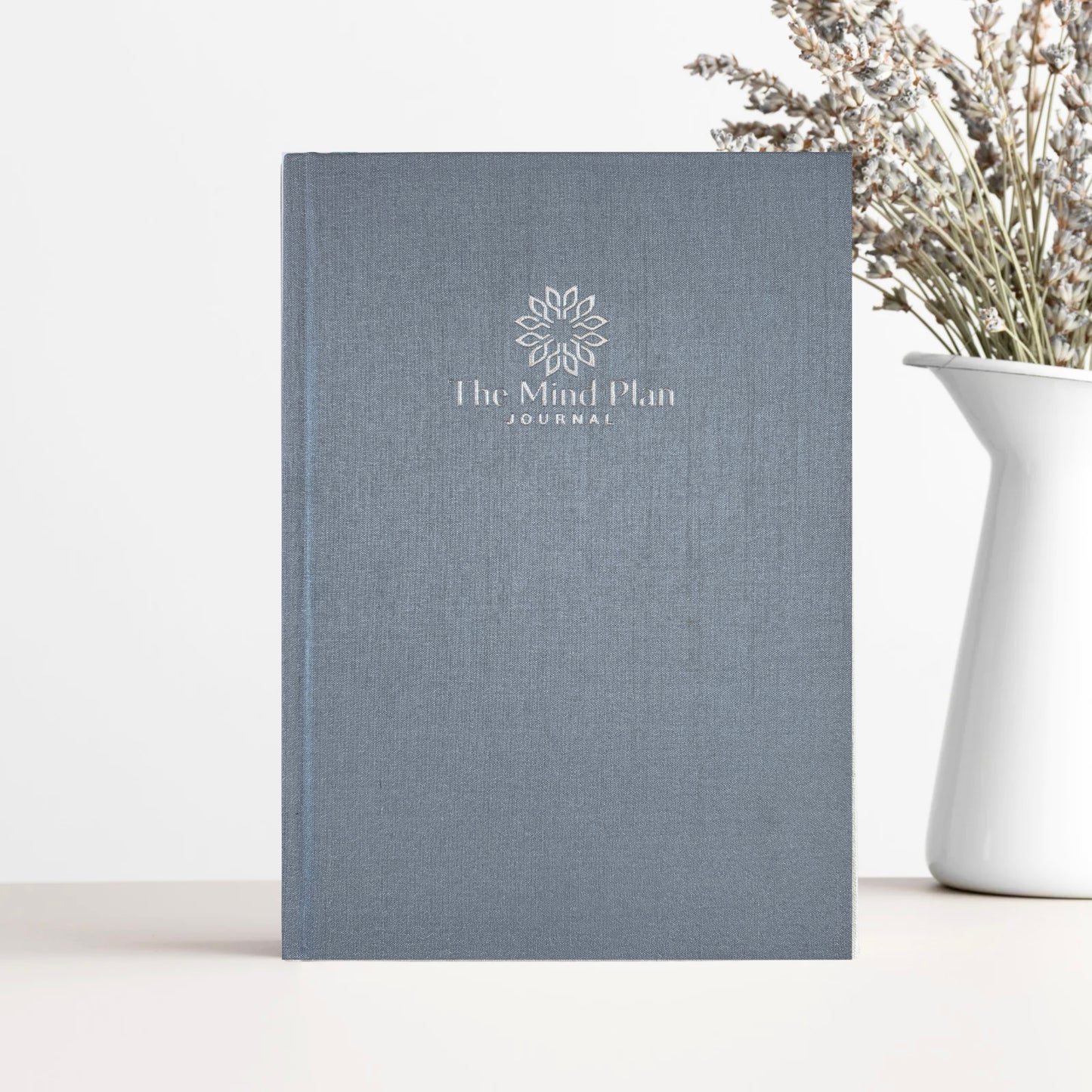 Gray journal titled 'The Mind Plan Journal' on a white surface with a vase of dried flowers.