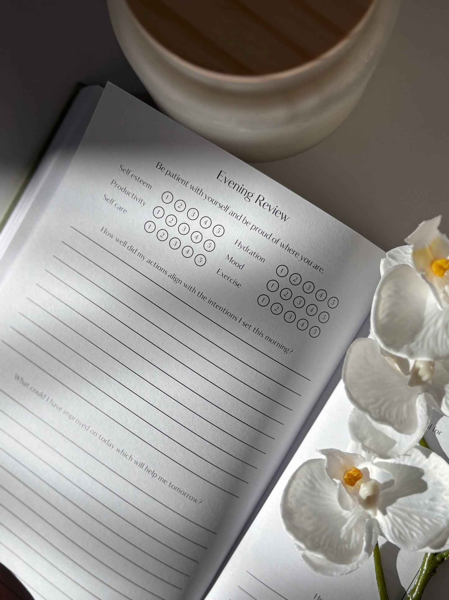 Journal with an evening review page, a cup, and white flowers on a light surface