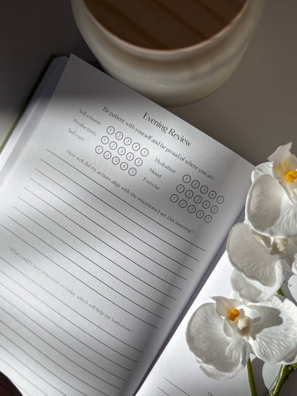 Journal with an evening review page, a cup, and white flowers on a light surface