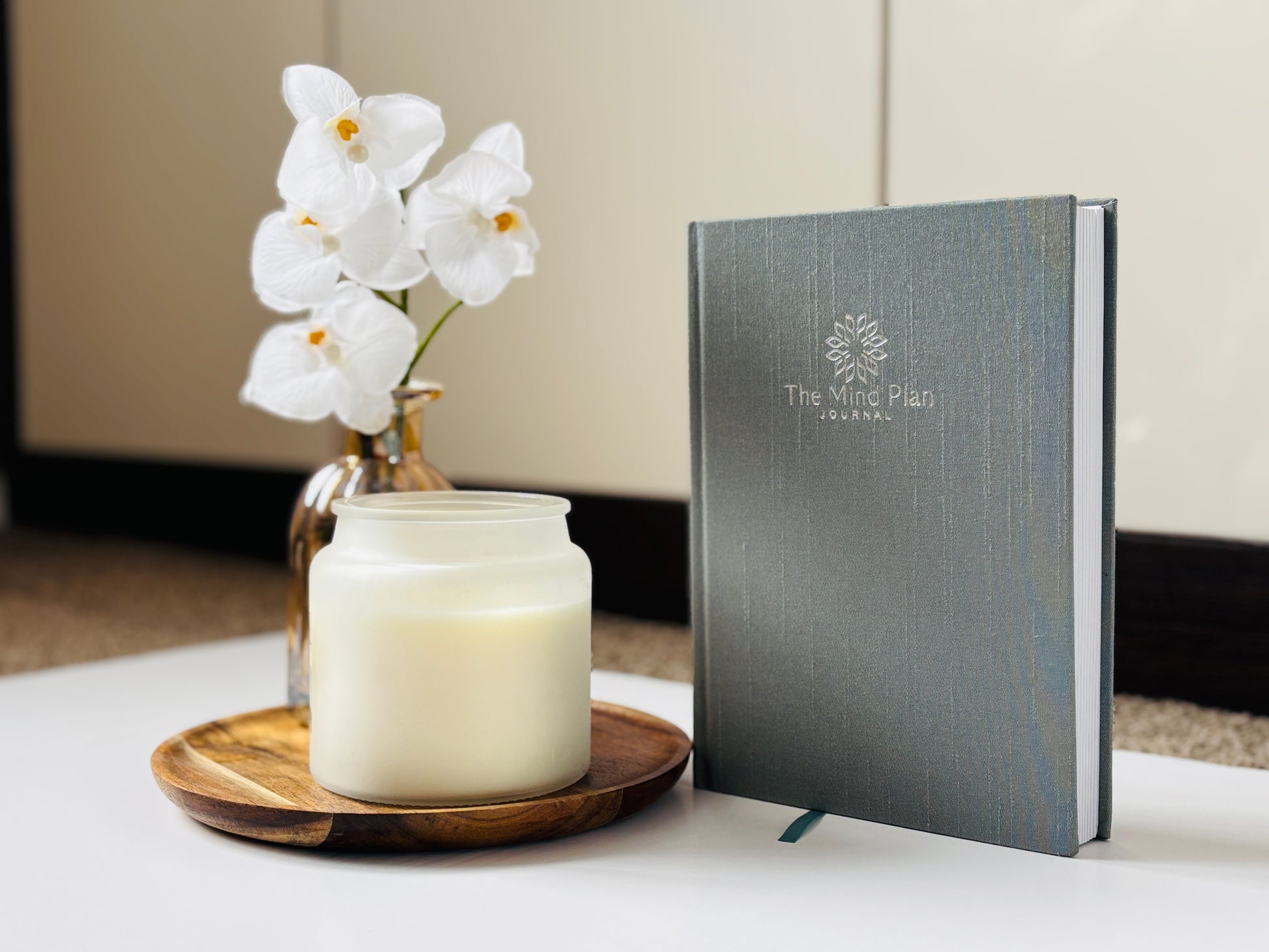 Blue Journal with 'The Mind Plan Journal' logo next to a candle and white flowers on a table.